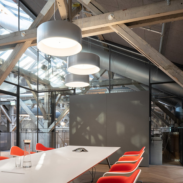 Three Artemide cylindrical pendant lamps in gray hang over a white conference table with orange chairs in an industrial loft space.