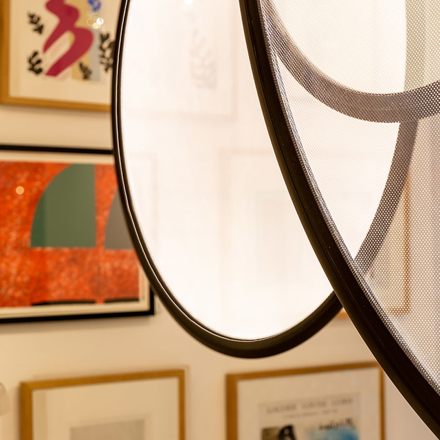 Close-up of a circular lamp with black metal frame and mesh diffuser, reflecting framed artworks on a gallery wall