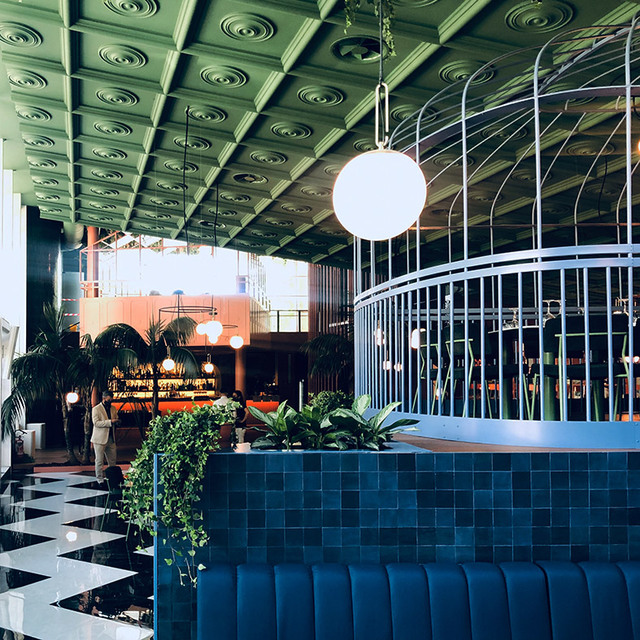 Modern restaurant interior with globe pendant lamp over blue tiled reception desk, ornate green ceiling, and tropical plants