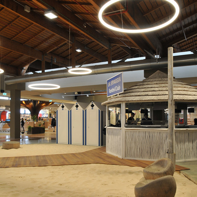 Indoor beach-themed space with circular pendant lights illuminating cabanas, sandy floor, and a thatched kiosk under a wooden ceiling.