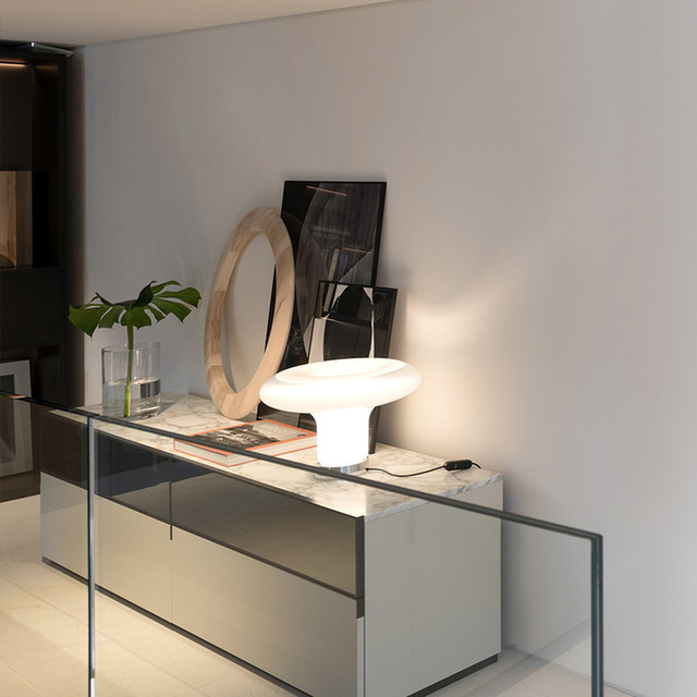 Artemide mushroom-shaped white glass table lamp glowing on a marble-top sideboard, styled with a round mirror and monstera plant