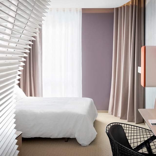 Modern hotel bedroom with white bed, mauve accent wall, taupe curtains, white venetian blinds, and a coral bedside lamp panel.