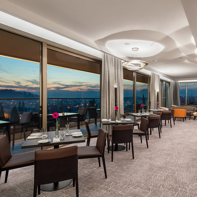 Elegant restaurant with panoramic city views at dusk, featuring a circular pendant lamp and warm cove lighting over dining tables.