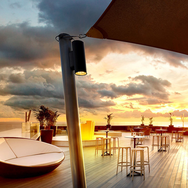Artemide outdoor cylindrical spotlight on a pole illuminating a rooftop bar terrace at sunset, with lounge seating and high tables