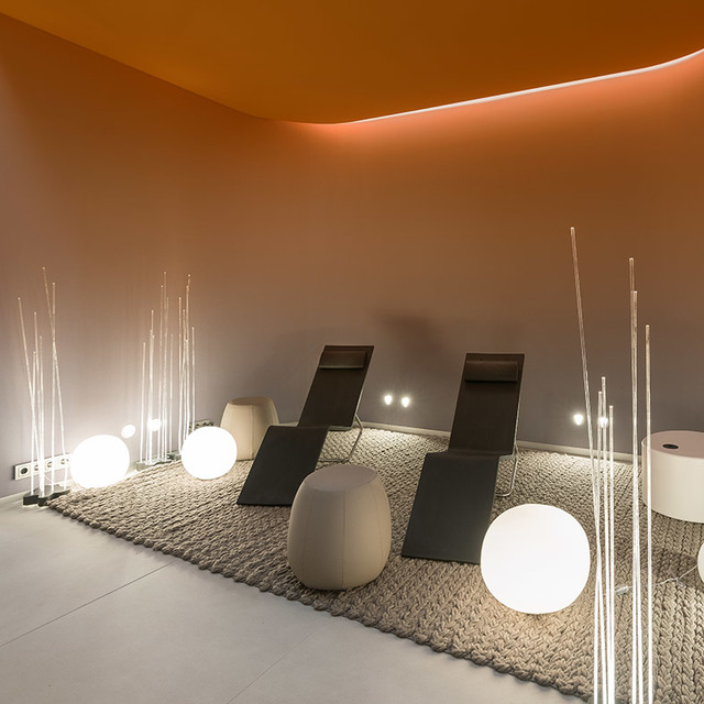 Relaxation area with glowing white spherical floor lamps and tall light sticks on a textured rug, beside dark reclining chairs under warm orange ceiling light