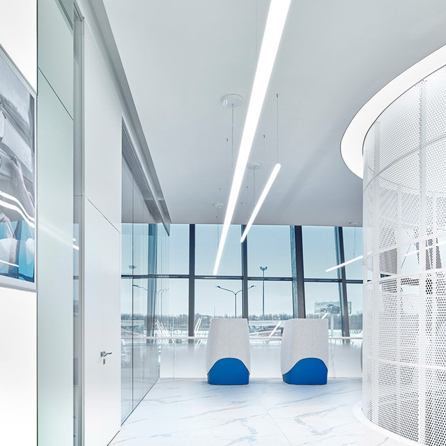 Modern office lobby with Artemide linear pendant lights on white ceiling, glass partitions, and two white chairs with blue seats.