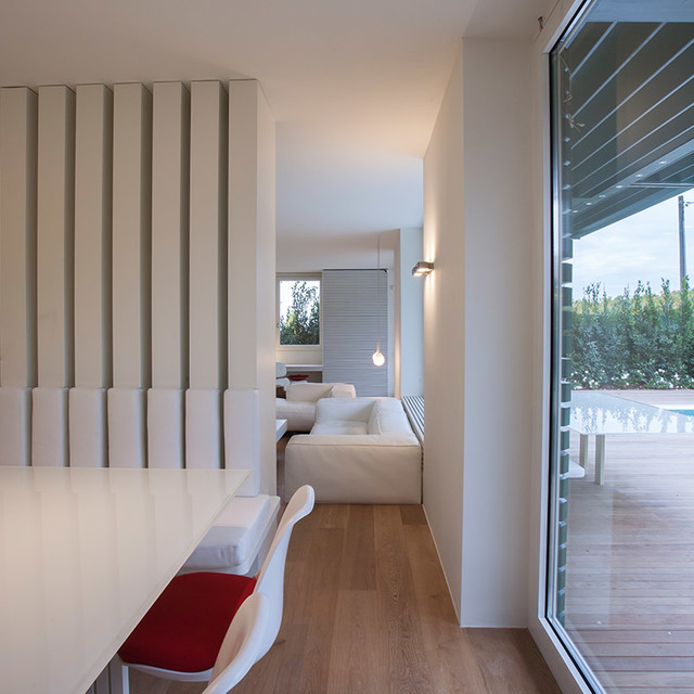 Modern living room with white walls, slatted partition, Artemide wall lamp, small pendant light, and dining area with red cushioned chair.