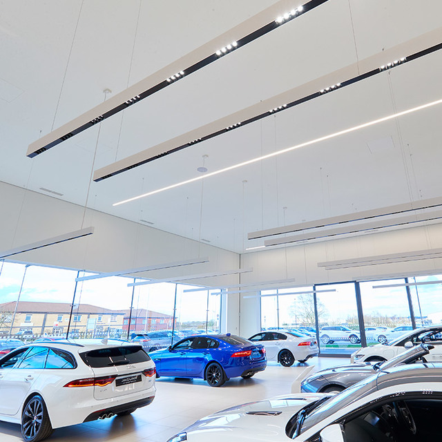 Car showroom with suspended linear LED lighting systems on white ceiling panels, illuminating luxury vehicles on display.