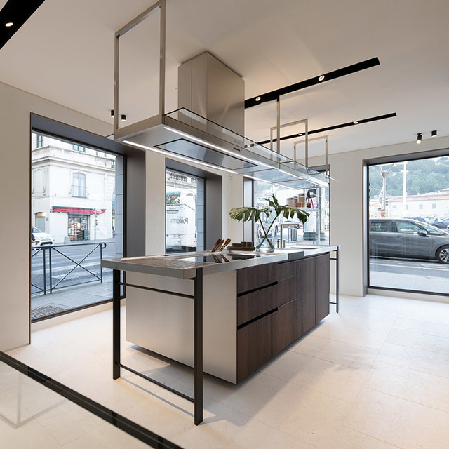 Modern kitchen showroom with central island, dark wood drawers, steel hood, and Artemide recessed spotlights on ceiling tracks.