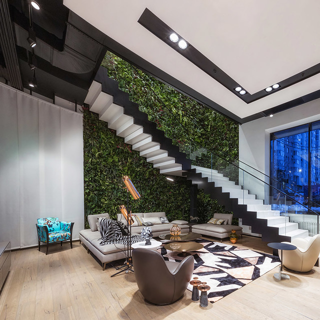 Modern double-height living room with green wall, floating staircase, track lighting on black ceiling beams, and contemporary furniture.