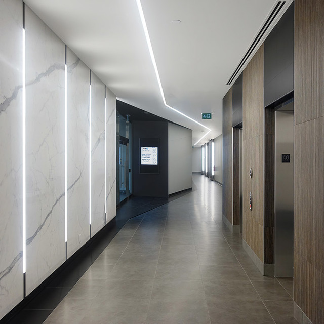 Modern corridor with recessed linear LED ceiling lights and vertical wall-mounted light strips illuminating marble panels near elevators.