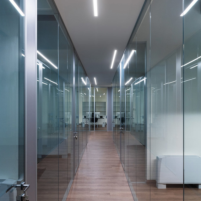 Modern office corridor with glass partitions and Artemide linear LED ceiling lights creating a bright, symmetrical perspective.
