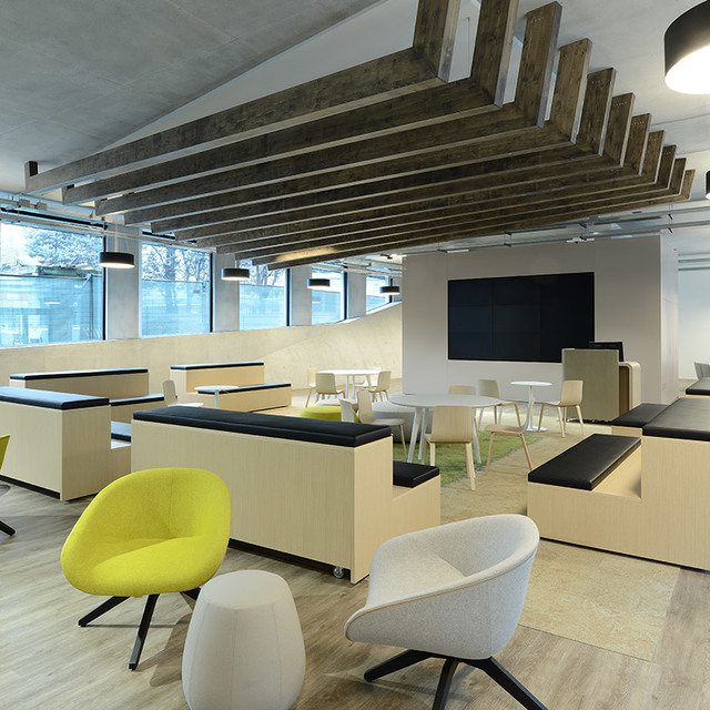 Modern office lounge with pendant lights, wooden ceiling beams, yellow and grey armchairs, booth seating, and a large display screen.