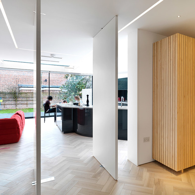 Modern open-plan kitchen with Artemide recessed linear LED ceiling lights, herringbone wood floor, glass partition, and garden view.