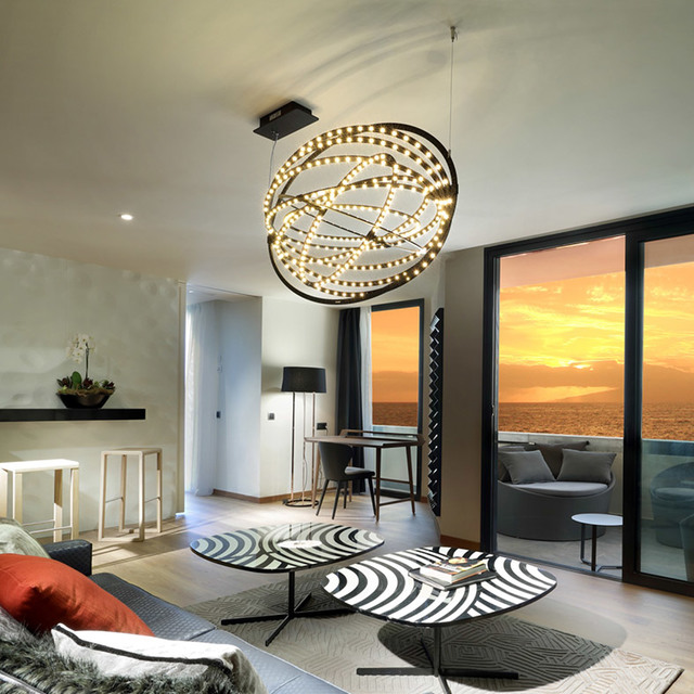 Spherical black pendant chandelier with intertwined LED rings illuminating a modern living room at sunset with ocean view.