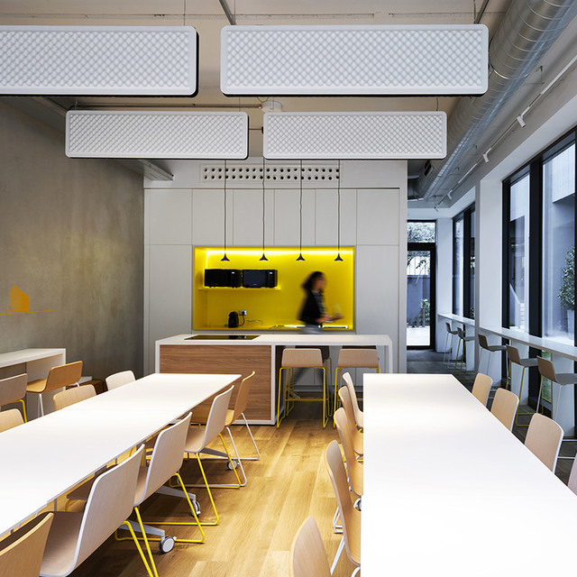 Modern office canteen with Artemide rectangular pendant lights illuminating white tables, chairs with yellow legs, and a yellow-accented kitchen area.