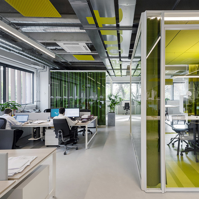 Modern open-plan office with linear ceiling lights, glass-partitioned meeting room, green accents, and employees working at white desks