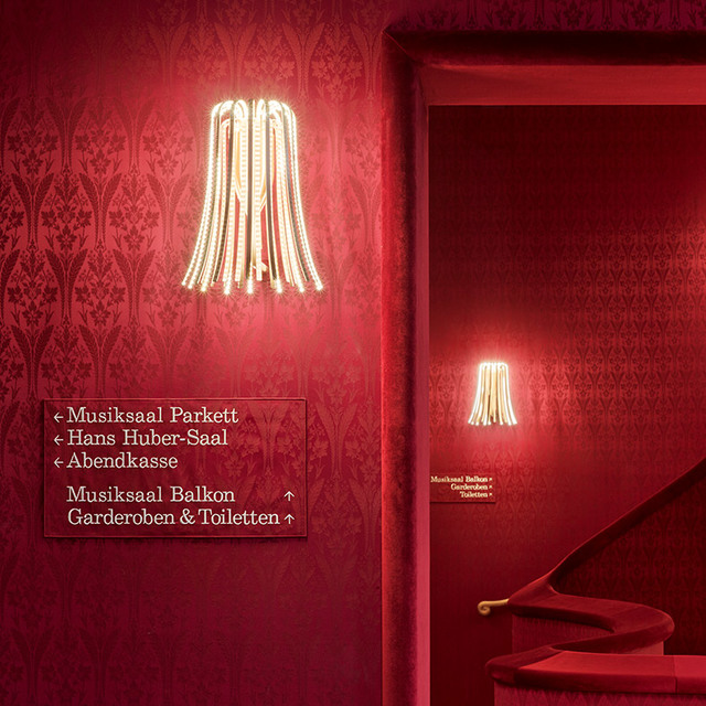 Artemide Calla wall sconces with cascading LED tubes illuminate a red damask corridor with directional signs in a concert hall.