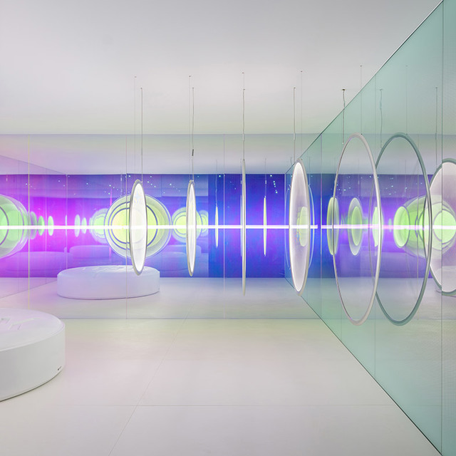 Futuristic white gallery with oval Artemide pendant lamps suspended on thin cables, reflected in glass walls amid vibrant purple and green light projections