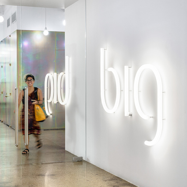 Artemide neon-style LED tube letters spelling 'big' mounted on a white wall in a modern office entrance, woman walking by.