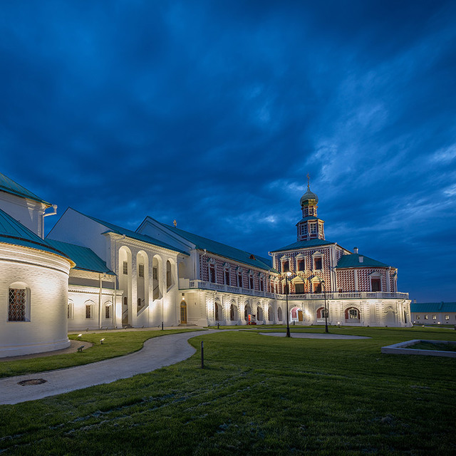 Historic Russian Orthodox monastery complex illuminated at dusk, with warm architectural lighting highlighting white walls and ornate bell tower