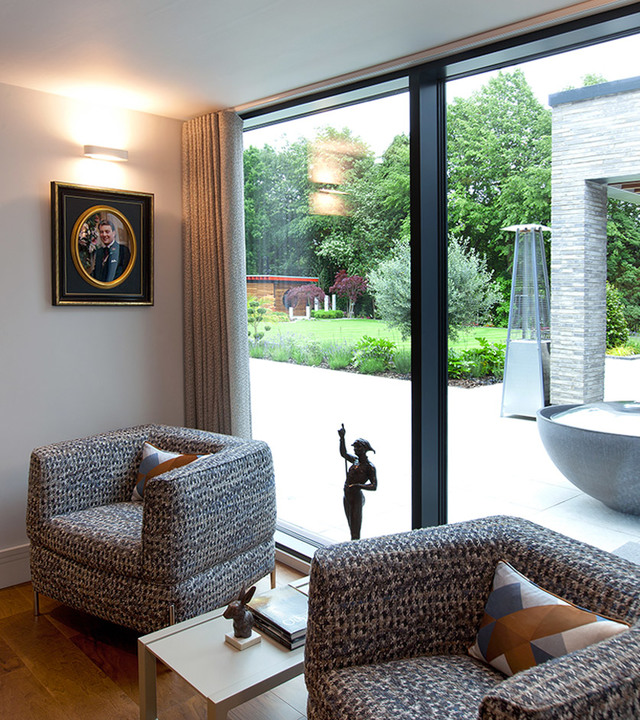 Modern living room with two patterned armchairs, a wall sconce providing warm light, large glass doors opening to a lush garden