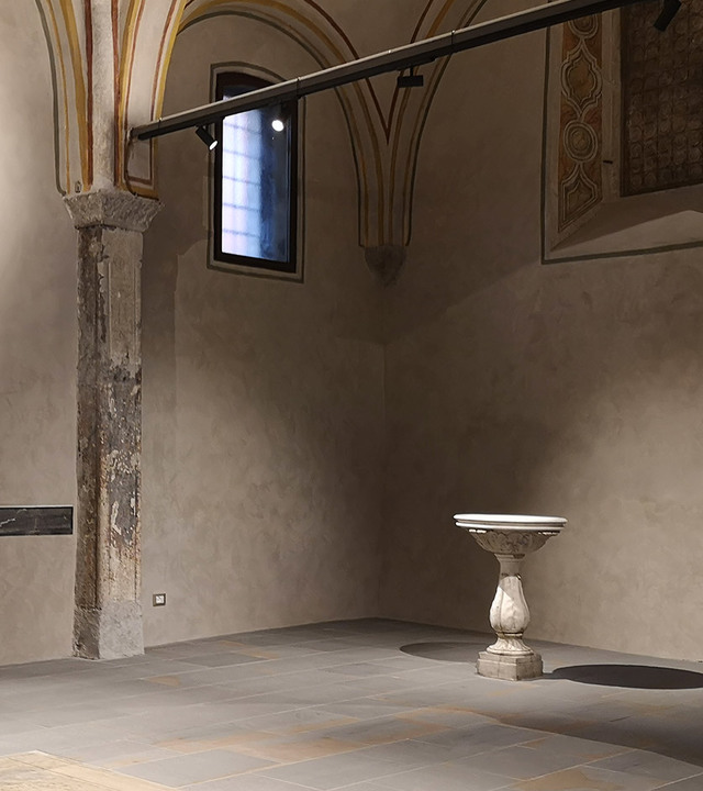 Historic vaulted interior with Artemide track spotlights illuminating a stone column and antique marble pedestal basin.