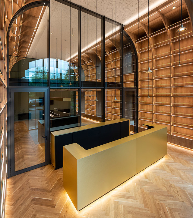 Modern reception area with gold desk, herringbone wood floor, arched wood-paneled walls, and warm linear LED ceiling lighting