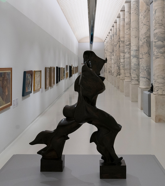 Museum gallery with a large dark bronze abstract sculpture, paintings on white walls, and marble columns under linear ceiling lighting.