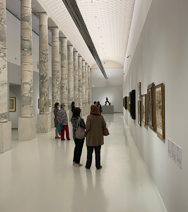 Museum gallery with marble columns, visitors viewing artwork on white walls, lit by soft indirect ceiling lighting.