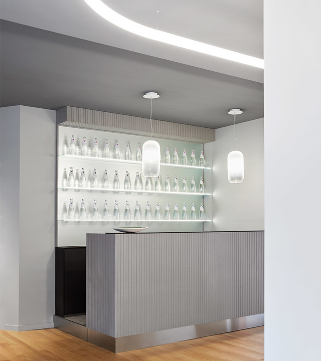 Modern bar counter with two white opal glass pendant lamps hanging from ceiling, backlit shelves displaying glass bottles.