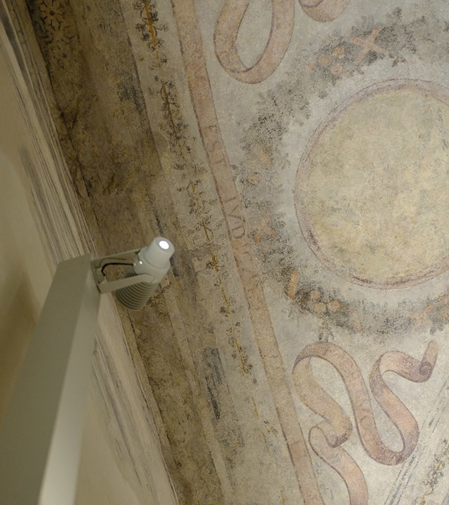 Artemide white adjustable spotlight mounted on wall, illuminating an ancient fresco with decorative motifs on a vaulted ceiling