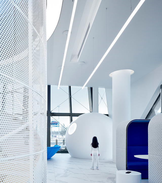 Futuristic white interior with linear ceiling lights, perforated wall panel, a spherical pod structure, and a person standing in the space