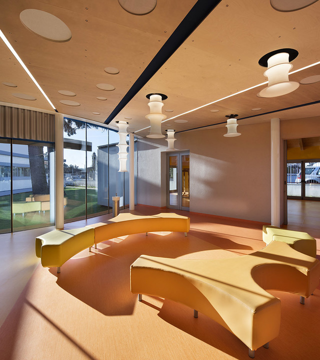 Modern lounge with curved yellow seating, warm terracotta floor, and white tiered ceiling pendant lamps in a bright open space