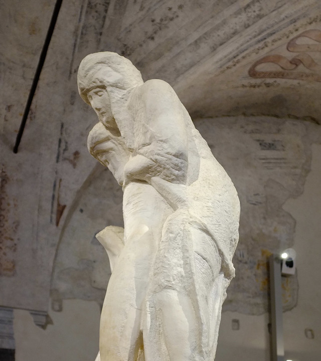 Michelangelo's unfinished Pietà Rondanini marble sculpture, showing a standing figure supporting a limp body, displayed in a museum.