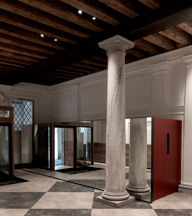 Historic interior with marble columns, wooden beam ceiling with recessed spotlights, checkered floor, and mirrored walls.