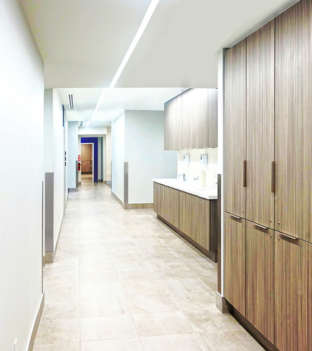 Modern office corridor with recessed linear LED lighting on white ceiling, wood-finish cabinetry, and neutral stone-tile flooring.