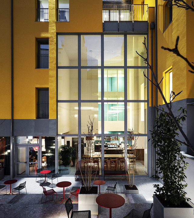Illuminated restaurant interior visible through large glass facade, with warm Artemide lighting, outdoor seating area with red tables and chairs.
