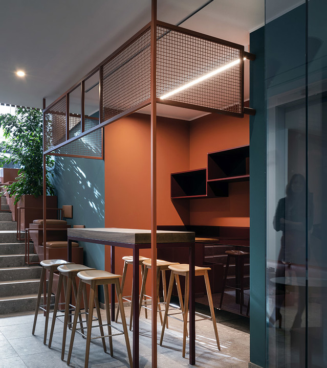 Bar area with high stools and a linear LED pendant lamp illuminating a mezzanine level with metal mesh railing and terracotta walls.