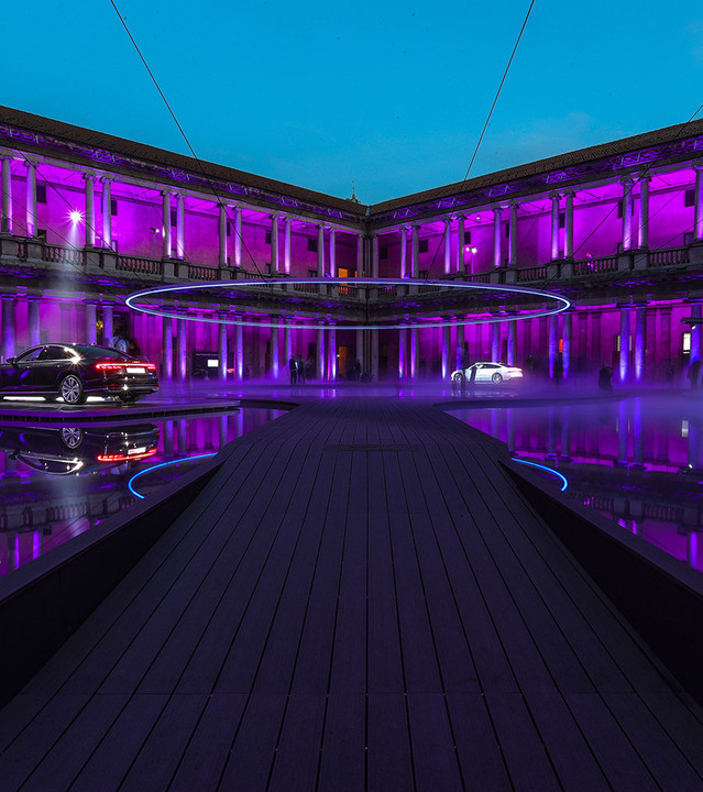 Neoclassical courtyard bathed in purple light with glowing circular neon ring installation, reflective floor, and two luxury cars on display