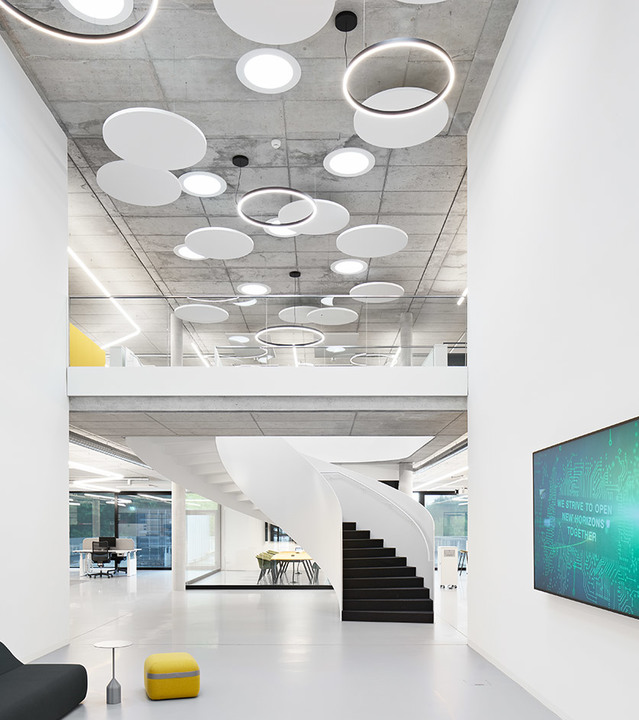 Modern office atrium with exposed concrete ceiling featuring circular and oval Artemide pendant lights, a spiral staircase, and open mezzanine.