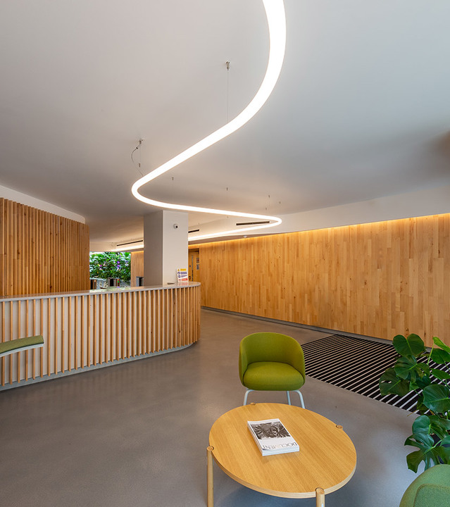 Modern lobby with a sinuous LED linear pendant light on the ceiling, wood-paneled walls, green armchair, and round wooden table