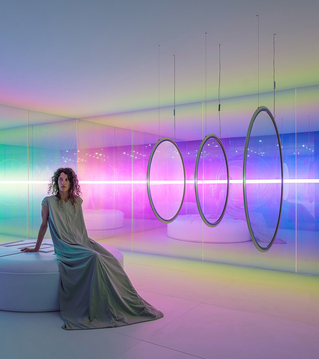 Woman in draped dress sits in immersive room with colorful RGB lighting, circular pendant rings, and vivid pink and green light beams