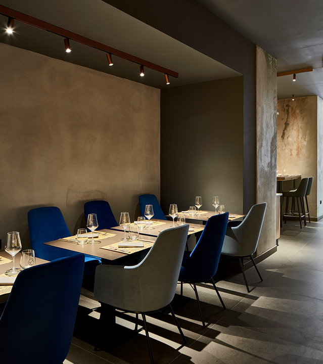 Elegant restaurant dining room with blue and grey velvet chairs, set tables, and Artemide track spotlights mounted on copper-colored rails.