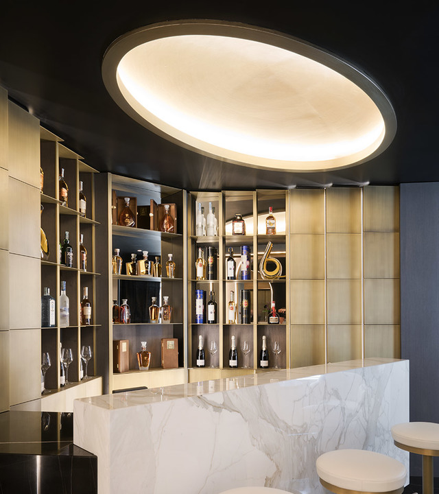 Elegant bar with large oval recessed ceiling light illuminating marble counter and gold-toned shelves displaying bottles.
