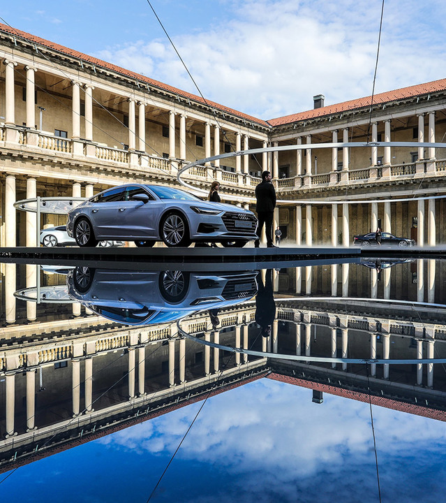 Audi sedan displayed on a raised platform in a classical colonnaded courtyard, reflected in a mirror-like water surface below.