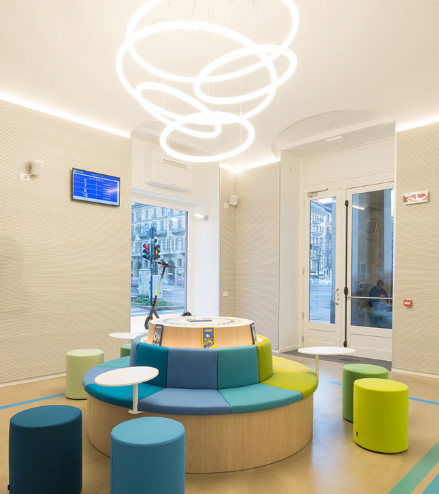 Modern waiting area with circular seating and colorful stools, lit by Artemide Overlap pendant lamp with interlocking luminous rings.