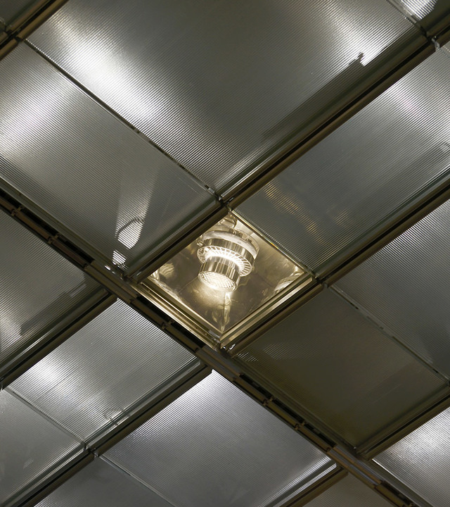 Recessed ceiling light fixture with metallic reflector and exposed bulb, set within a grid of translucent glass panels.