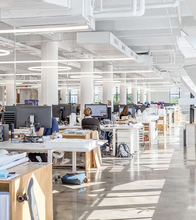 Open-plan office with workstations under circular and linear pendant LED lights on a white ceiling with exposed ducts.