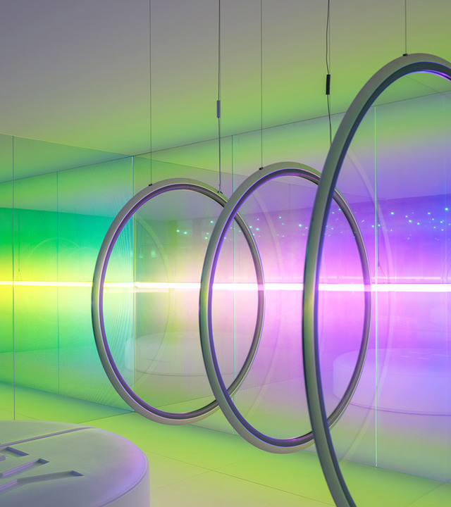Large circular pendant rings emit vibrant green, purple, and white light, creating an immersive chromatic atmosphere in a mirrored room.